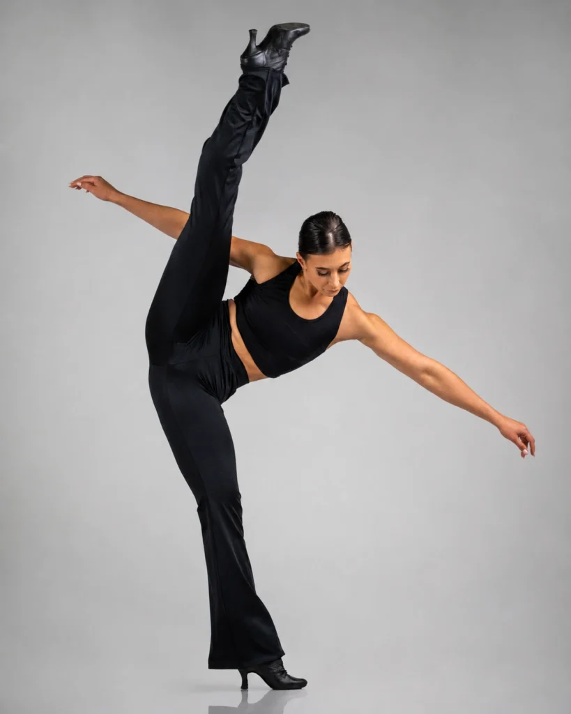 Dancer in black performing a high-leg pose against a light grey studio background.